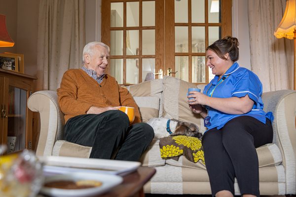 A Male Customer and Female Bluebird Care Assistant Enjoy A Hot Drink Together On A Sofa