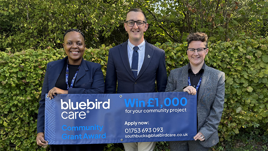 Comm Grant Launch 2025 - BUR Bluebird Care staff stand in front of a banner which announces the launch of a £1,000 community grant, which will be awarded to fund a project that benefits the local area.