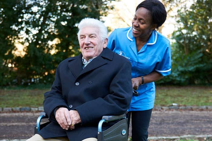 A Bluebird carer smiles while pushing an elderly gentleman in a wheelchair
